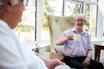 White man talking to an elderly man sitting drinking a cup of tea